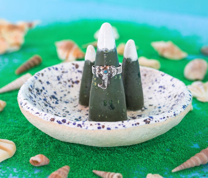 Silver ring with blue stone on ceramic ring holder and artificial green sand with seashells on a blue background